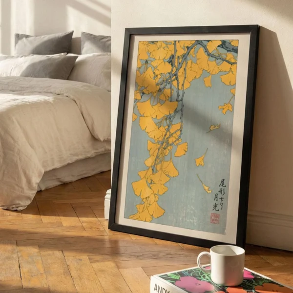 May include: A framed art print featuring a branch of yellow ginkgo leaves against a light blue background. The print has Japanese text in the lower right corner and is in a black frame. A white mug sits on a book in front of the print.